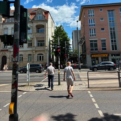Foto von einer Straße, über welche zwei Fußgänger gehen. Zu sehen ist eine Fußgängerampel auf der anderen Seite der Straße, welche jedoch rot anzeigt.